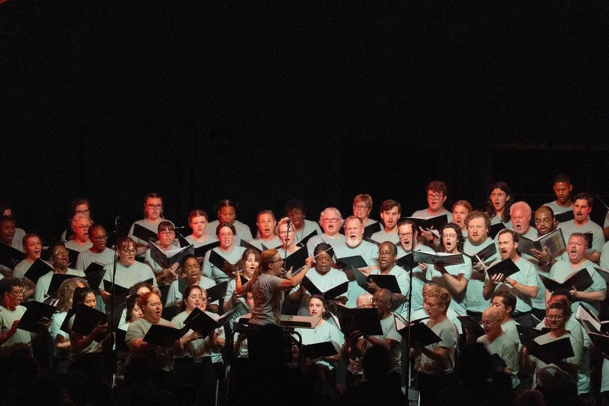A choir of men and women singing, conducted by a woman using hand gestures, on a dark stage.