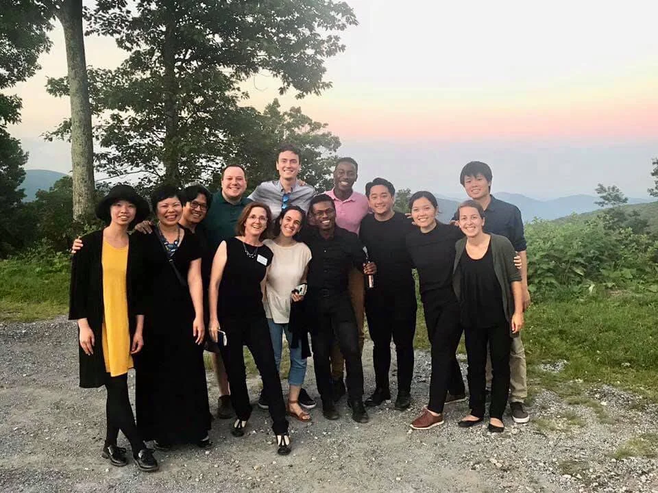 A group of twelve diverse friends standing outdoors on a gravel path with trees and mountains in the background during sunset, smiling and enjoying each other's company.