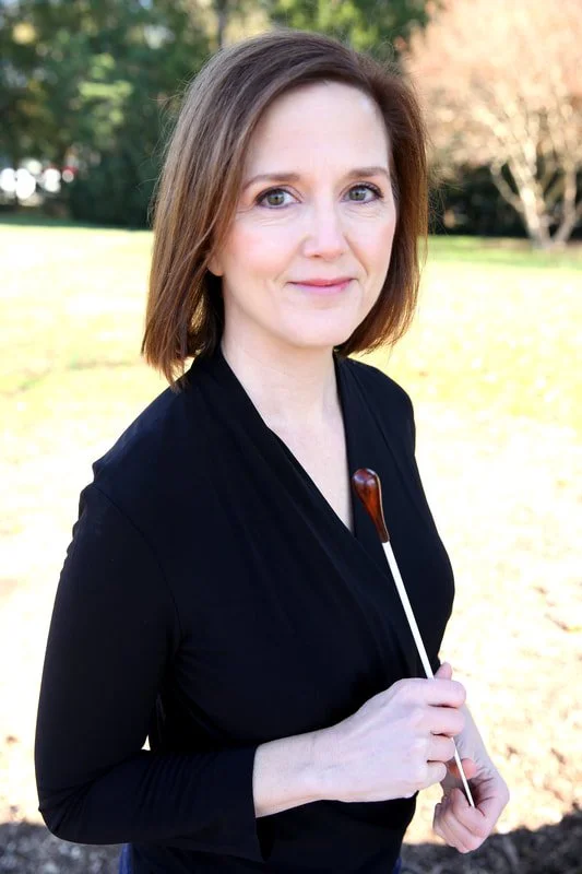 Erin Freeman wearing a black top, standing outdoors in a park with trees and grass in the background, holding a conducting baton.