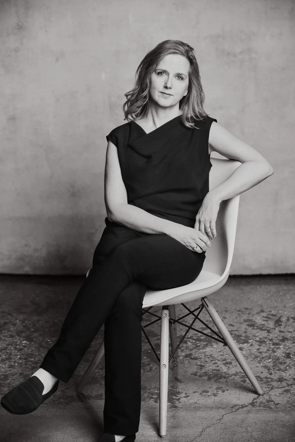 Erin Freeman wearing a sleeveless dark top and dark pants, sitting on a modern chair against a plain background.