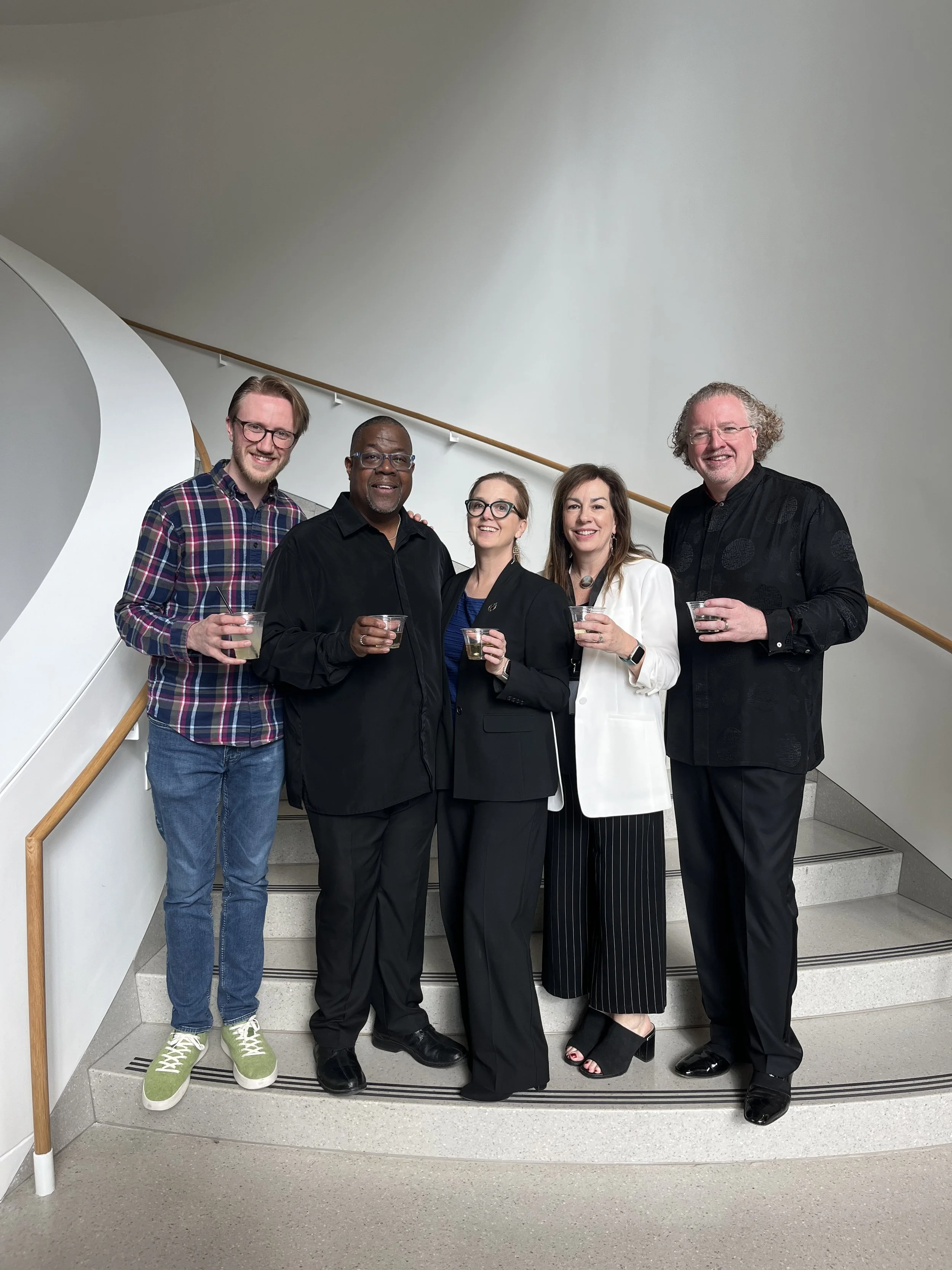 Group of five diverse professionals standing on a staircase, holding drinks and smiling at the camera, in a modern indoor setting.