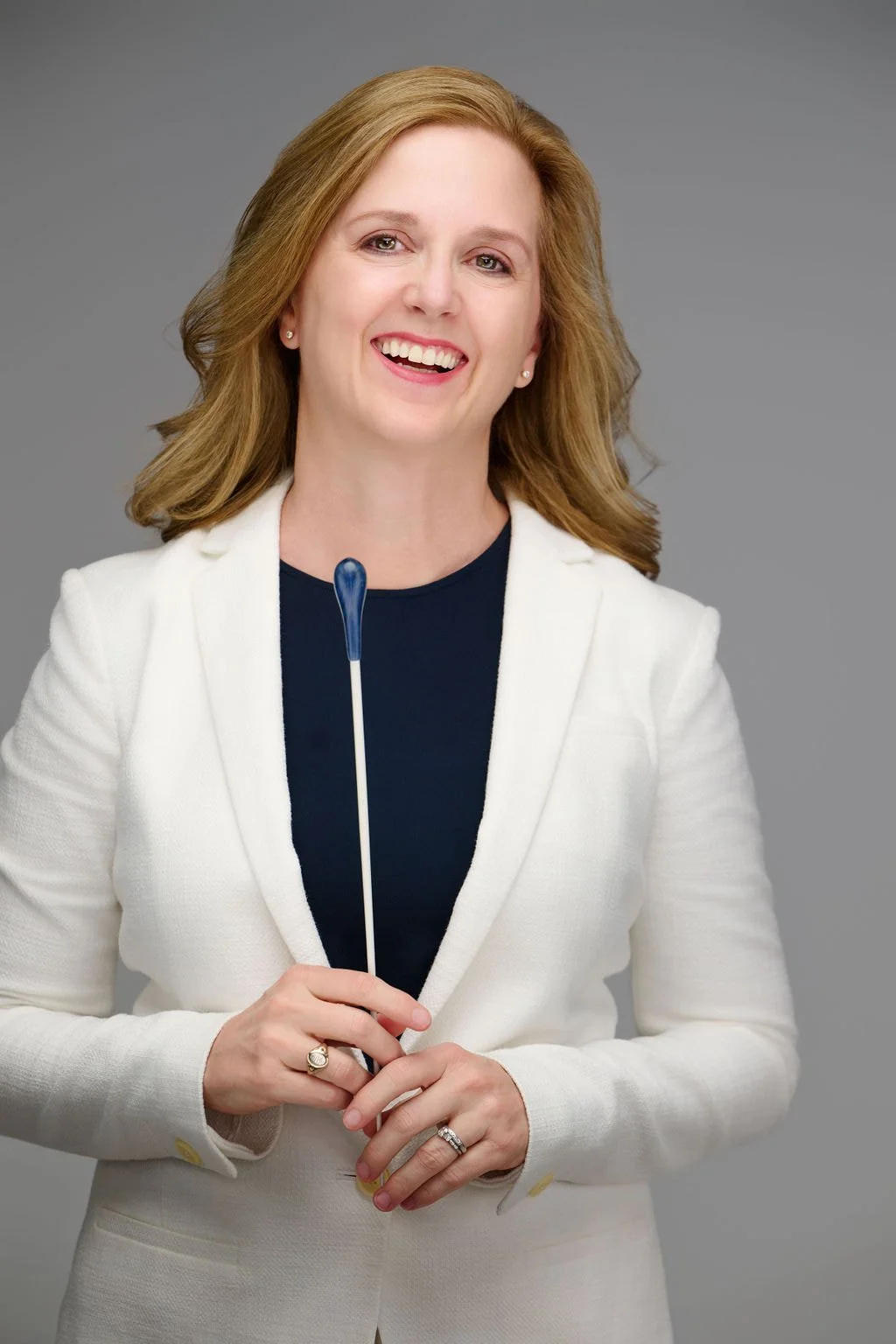 A woman with light brown hair smiling, wearing a white blazer and holding a blue and white pin with both hands, standing against a gray background.