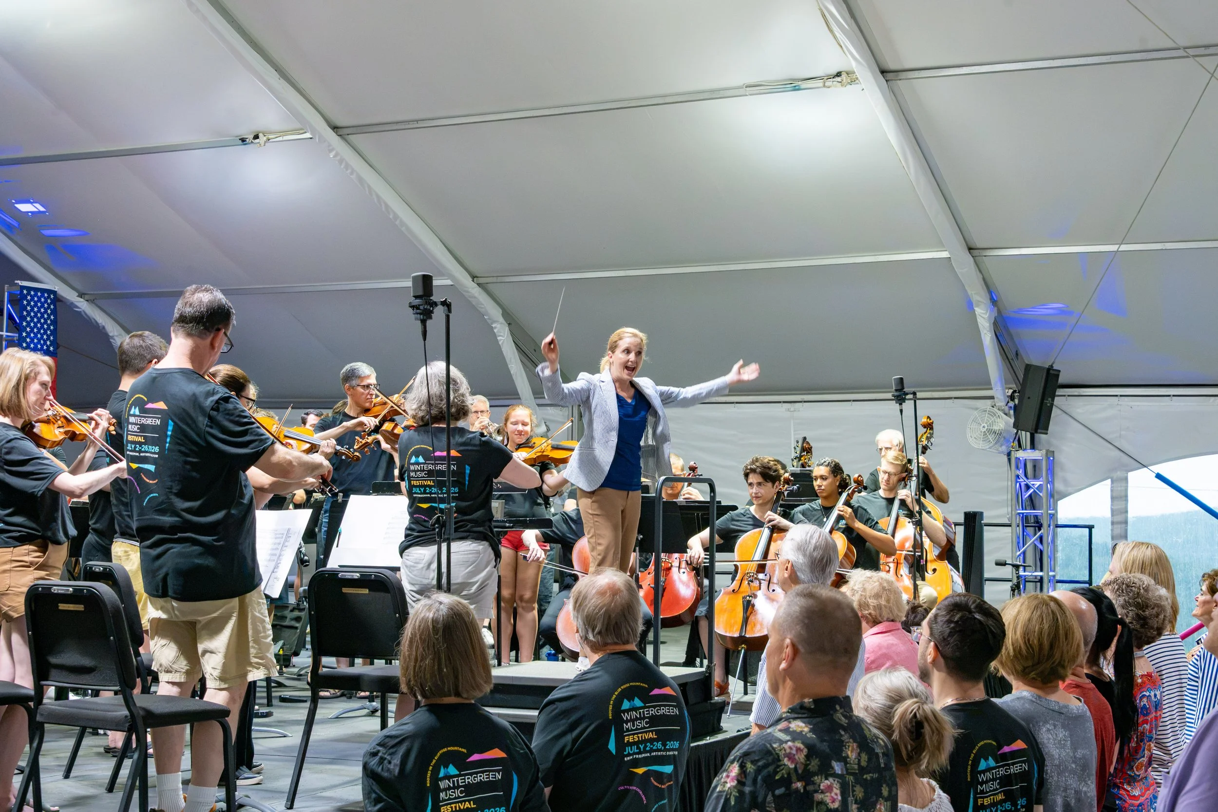 Orchestra performing under a large white tent, with a Erin Freeman conductor leading the musicians, an audience watching, and festival signage reading 'Wintergreen Music Festival July 2-26, 2026'.