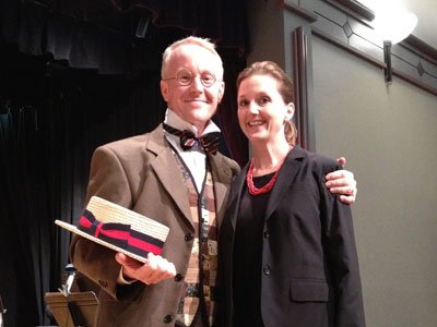 An elderly man and a middle-aged woman smiling and posing together on a stage, with the man holding a pair of shoes and the woman with her arm around him.