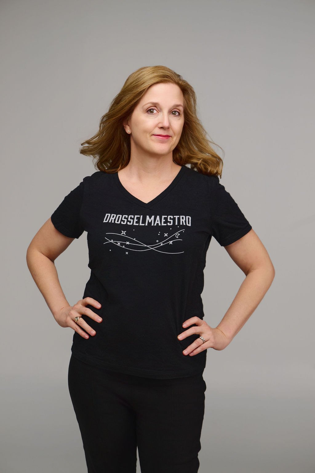 Erin Freeman with hands on her hips, wearing a black T-shirt with the word 'DROSSELMAESTRO' and a starry line design, against a plain gray background.