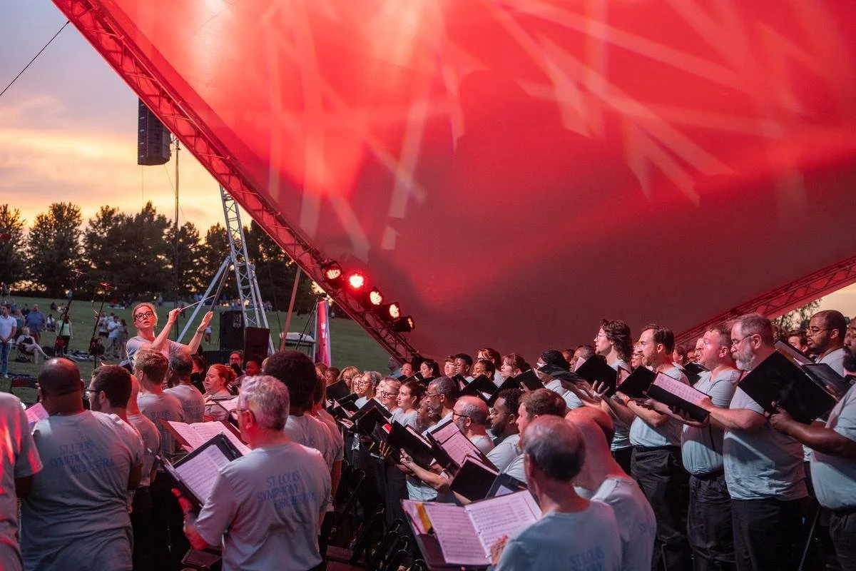 A large choir concert outdoors at sunset under a red canopy with a conductor leading the choir.