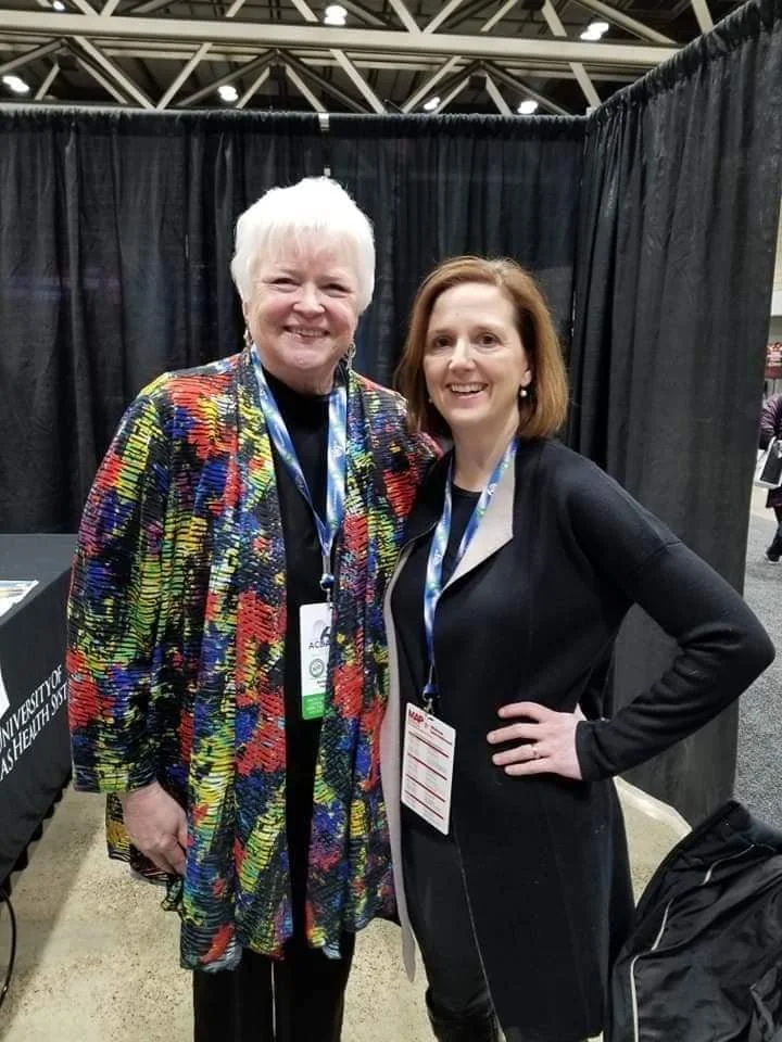 Two women smiling and posing at a conference or event with black curtains in the background. One woman has short white hair and is wearing a colorful patterned jacket. The other woman has shoulder-length reddish hair and is dressed in a black blazer 