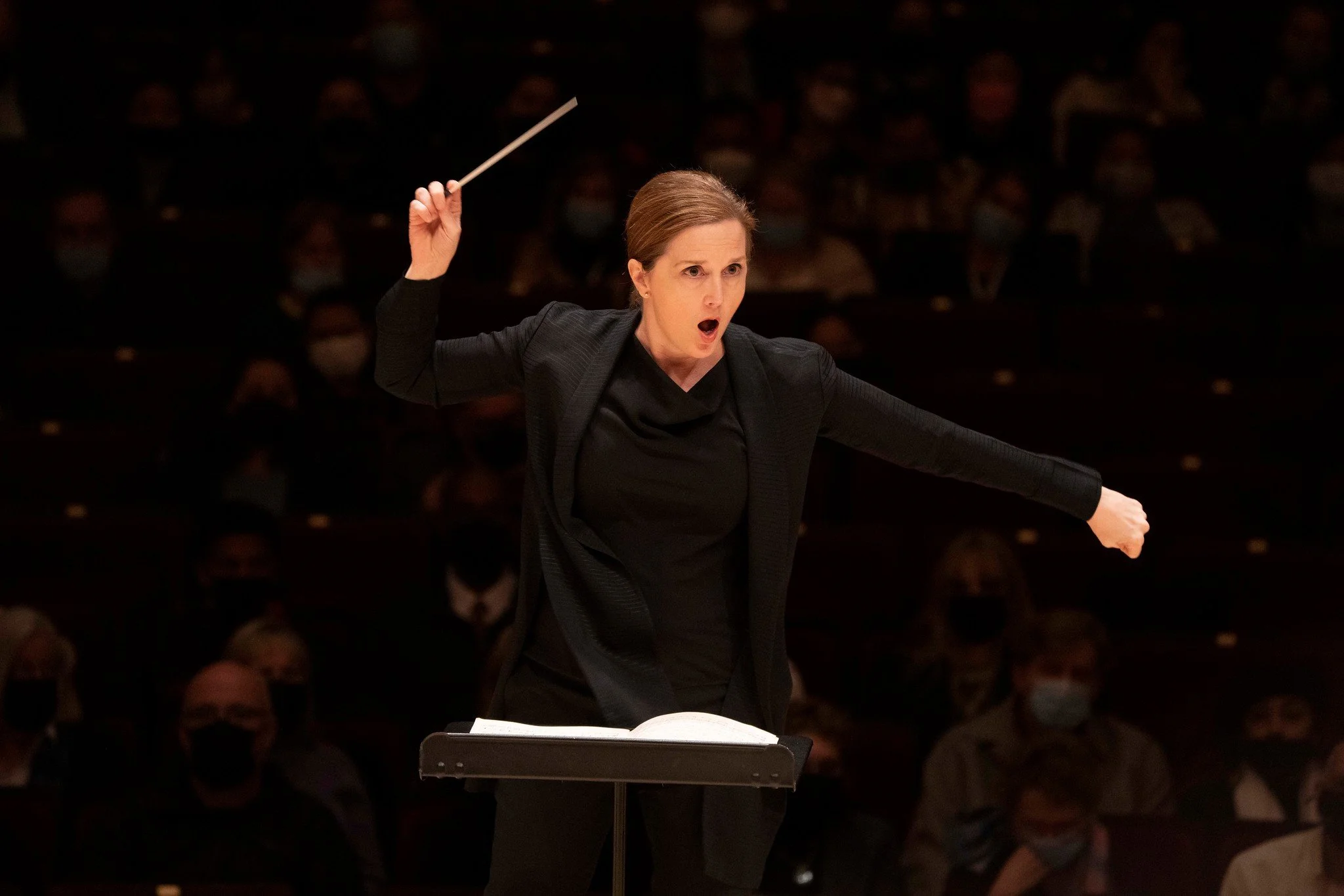 A female orchestra conductor passionately leads an orchestra during a performance, standing at a podium with music sheets, with an engaged audience in the background.