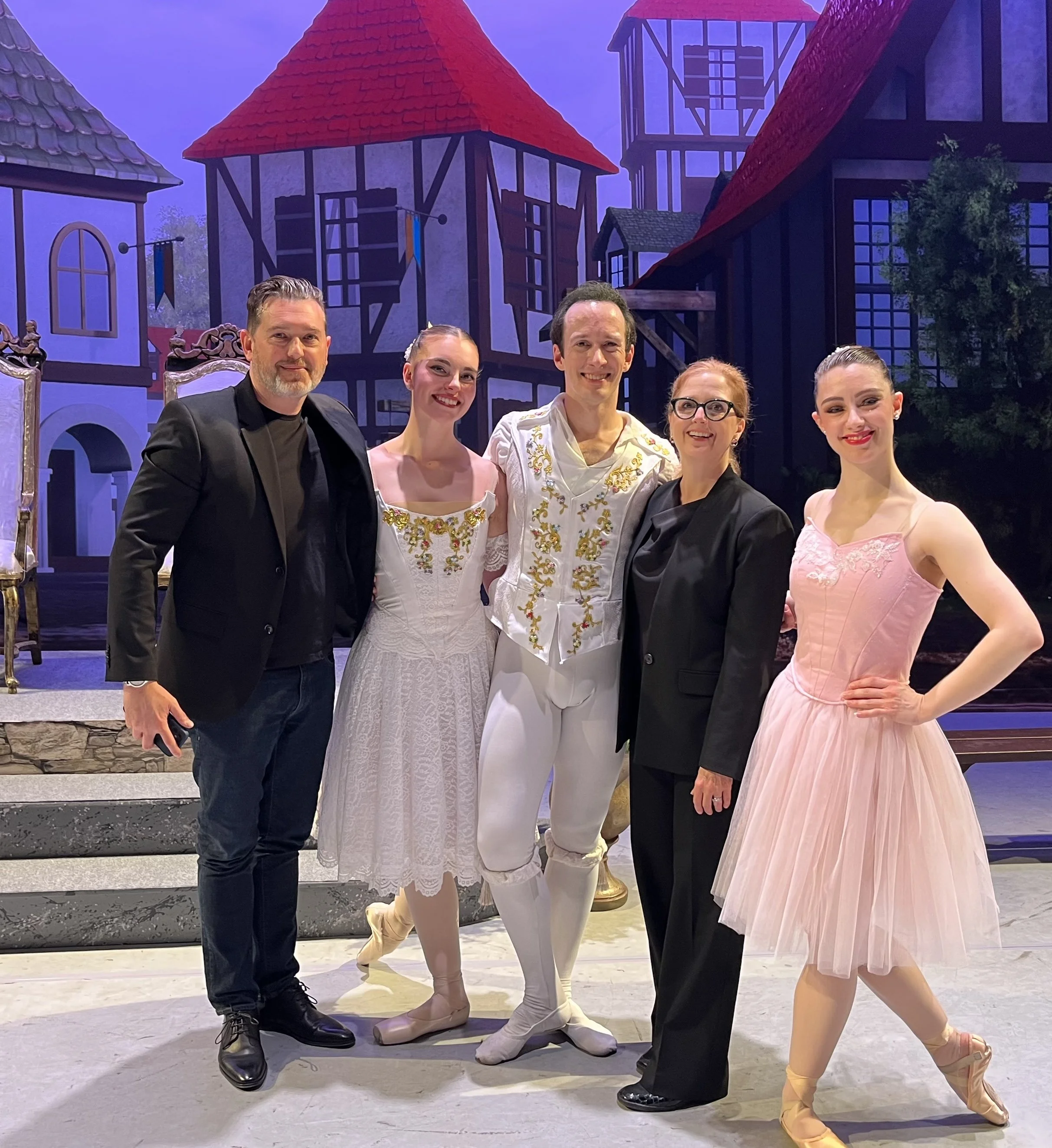 Five ballet dancers standing on stage in front of a painted backdrop of a whimsical village scene, dressed in ballet costumes, with one man in black formal attire. The dancers are smiling and posing for the photo.