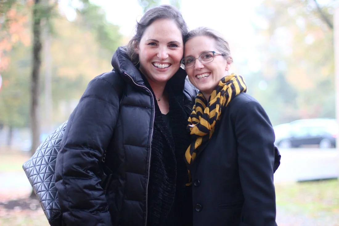 Two women smiling outdoors, dressed warmly for fall, standing close together in a park with autumn trees in the background.