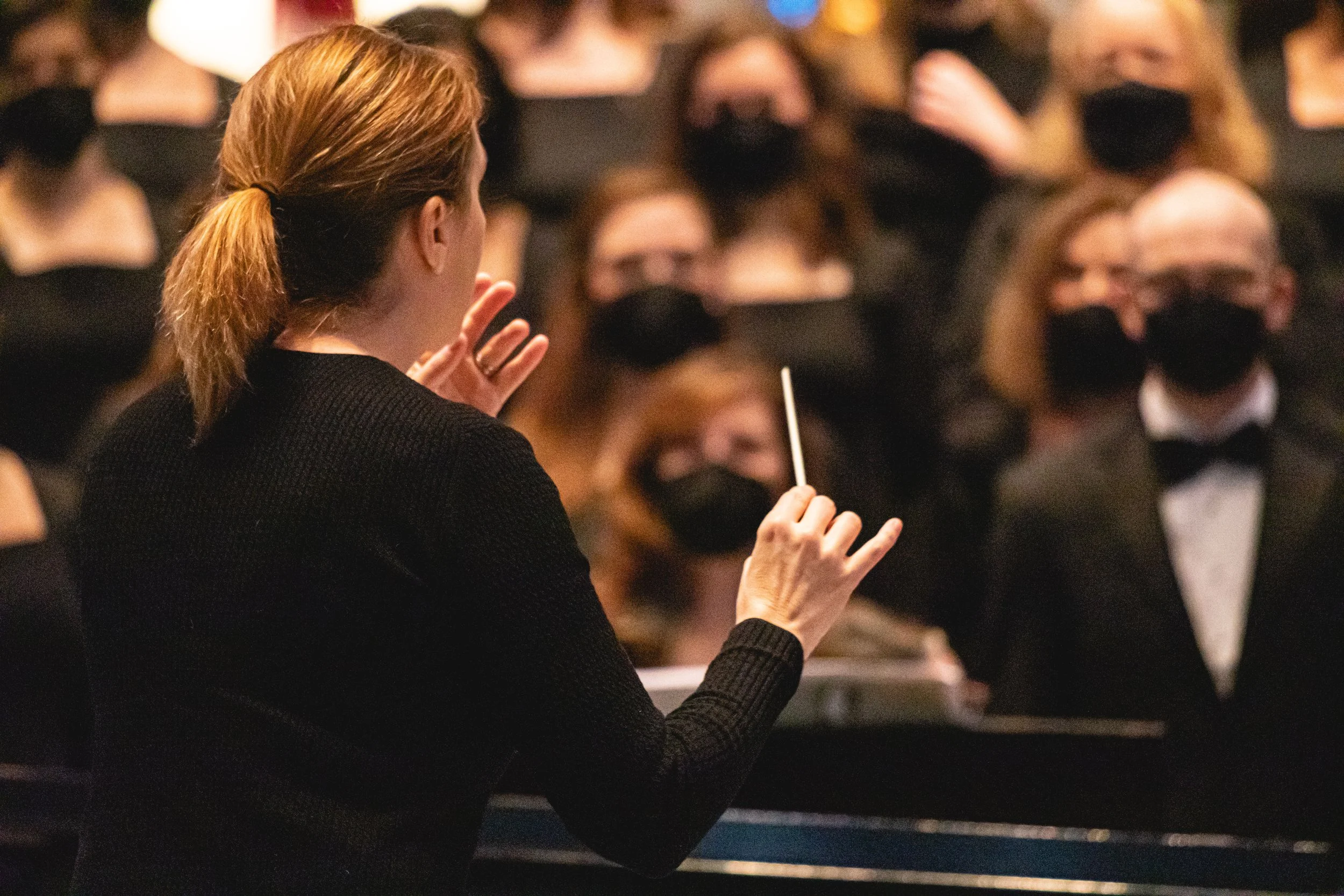 A woman with brown hair tied back is conducting or giving a speech in front of an audience wearing black face masks and black attire, holding a baton or pointer.