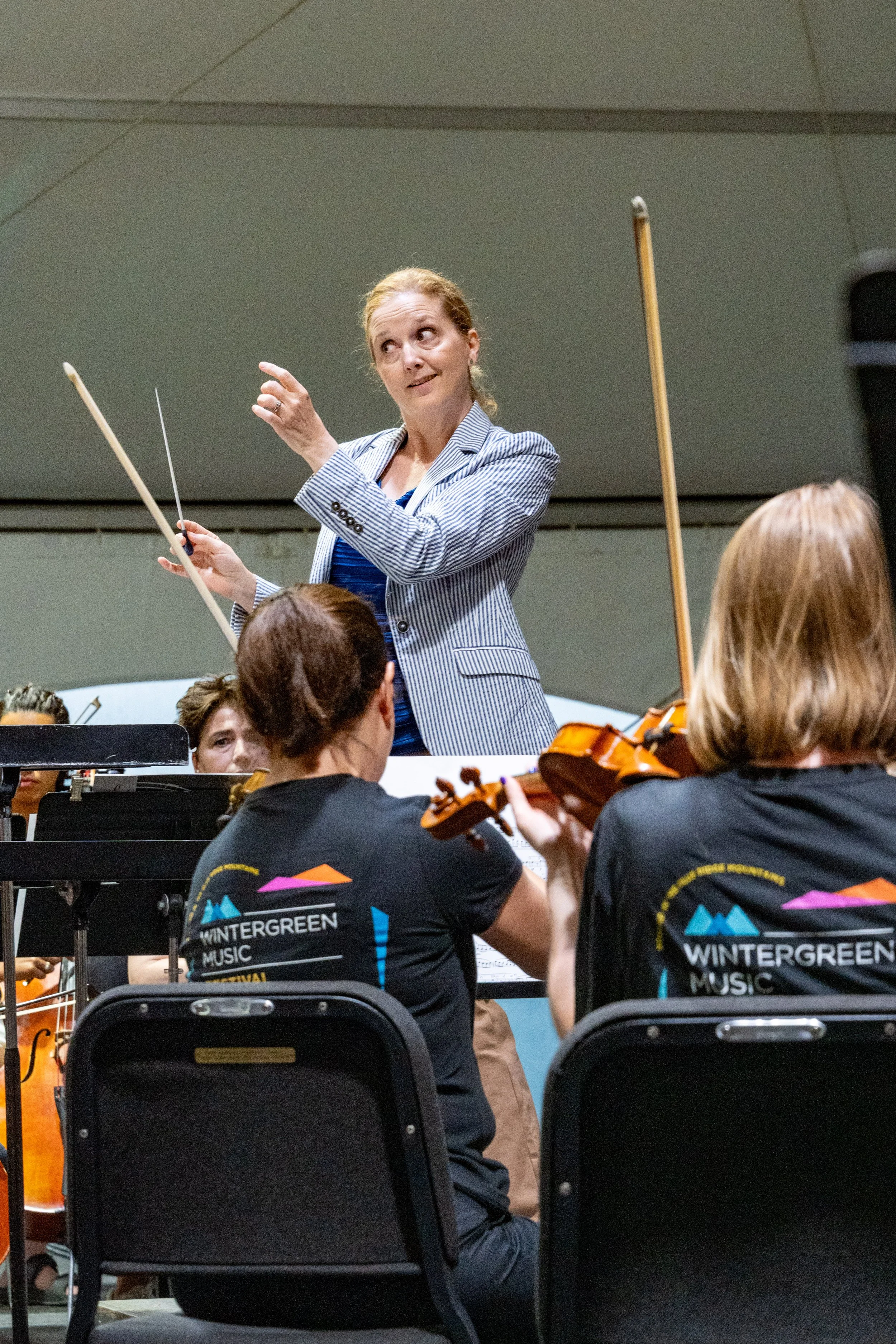 Erin Freeman leading an orchestra in a performance, with musicians in black shirts with 'Wintergreen Music Festival' logos on the back, playing string instruments. 