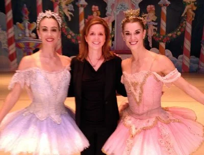 Three women posing together in costumes, with a colorful scene in the background that appears to be from a ballet or performance.