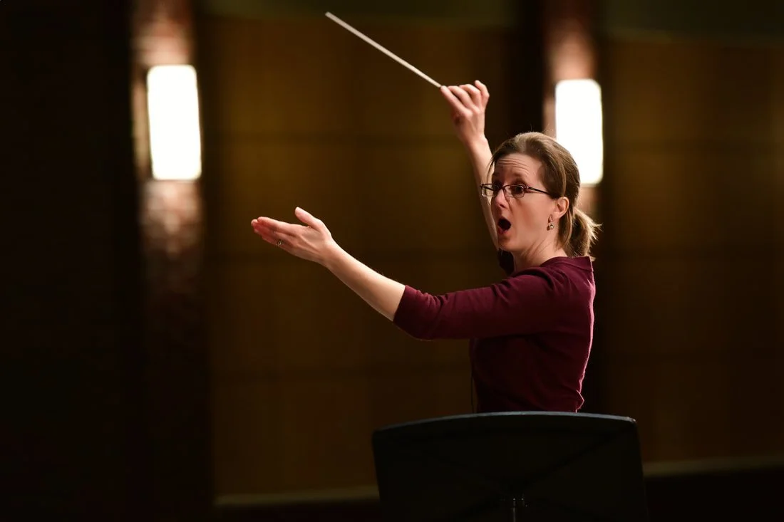 A woman conducting an orchestra, holding a baton in her right hand with a surprised or intense expression.
