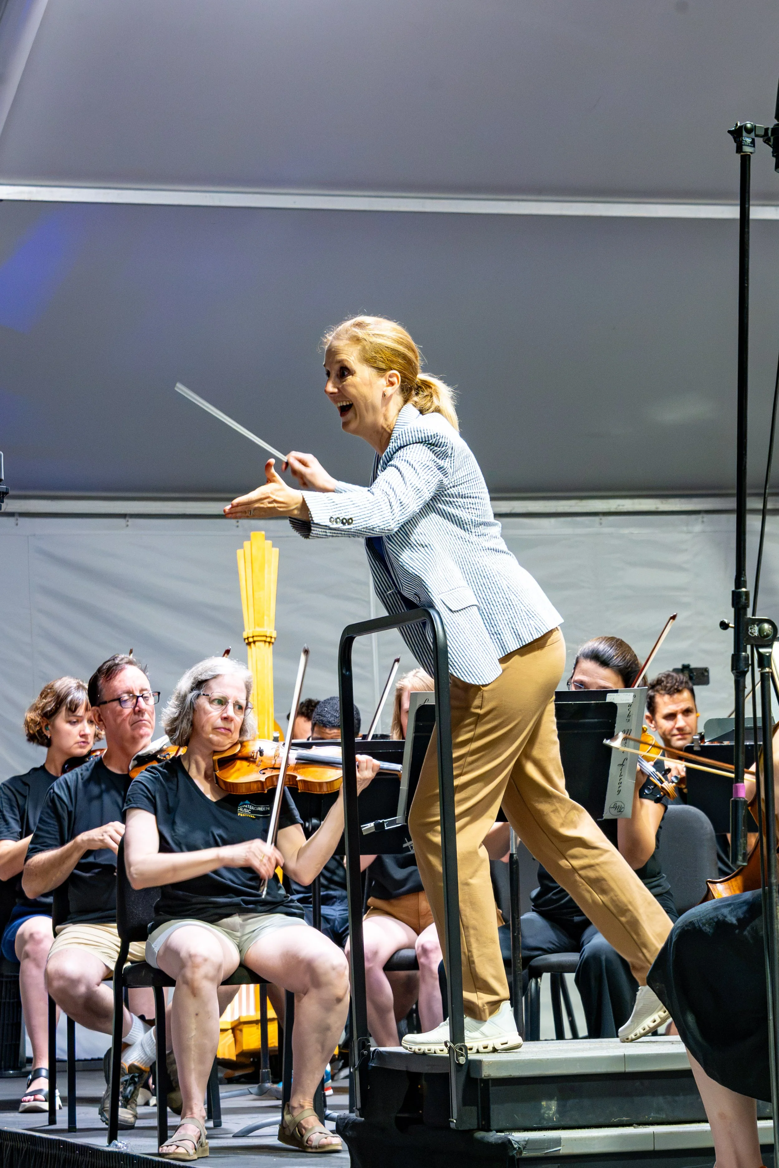 A woman in a striped blazer and beige pants conducting an orchestra on stage. She is standing on a chair, gesturing energetically with her hands, and appears to be smiling or singing. The orchestra members, seated with their music stands, are playing