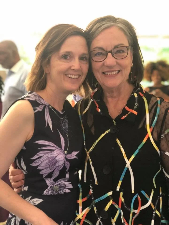 Two women smiling and standing close together at a social event, with blurred people in the background.