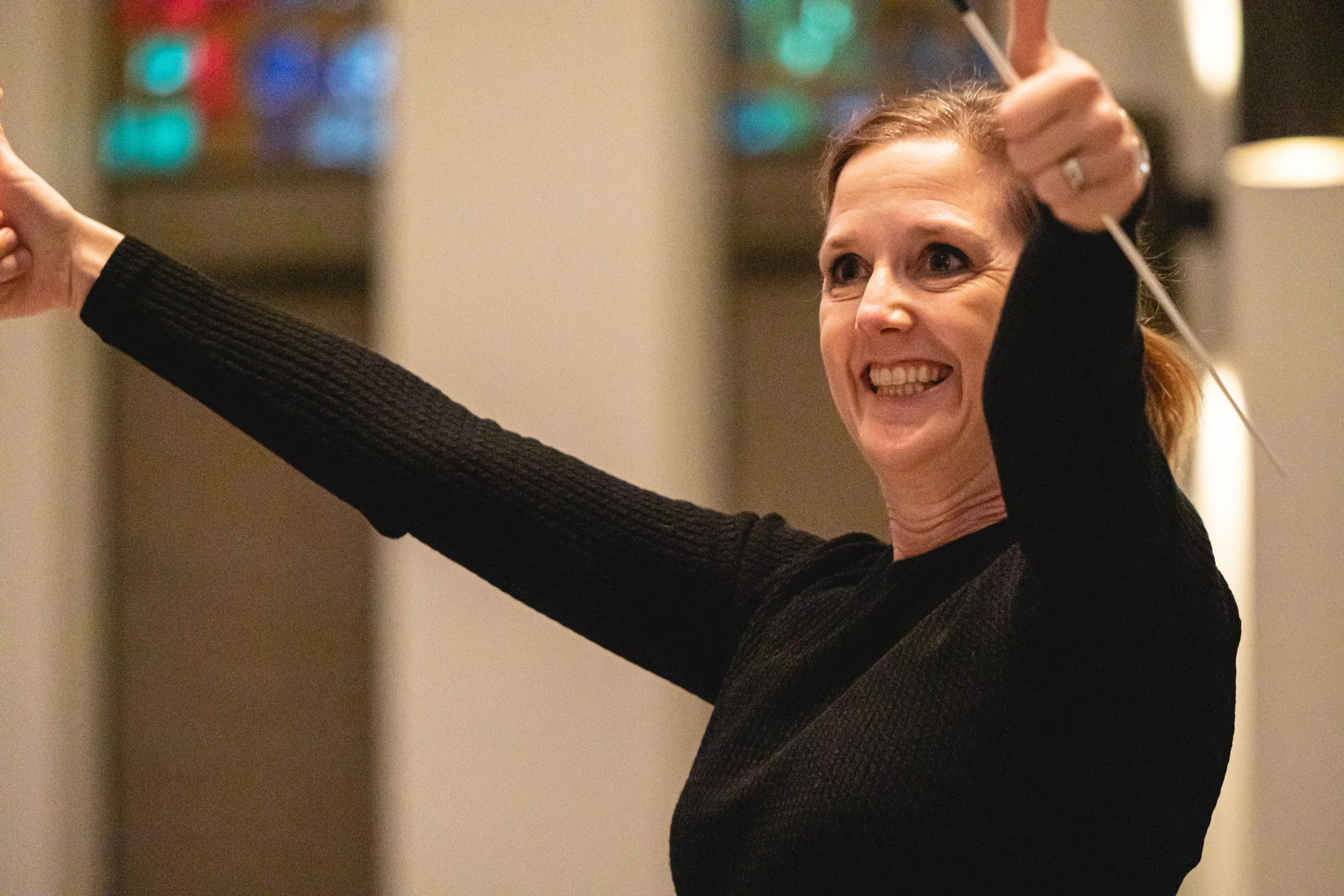 A smiling woman in black attire stretching her arms while holding a white object, in an indoor setting with a warm background.