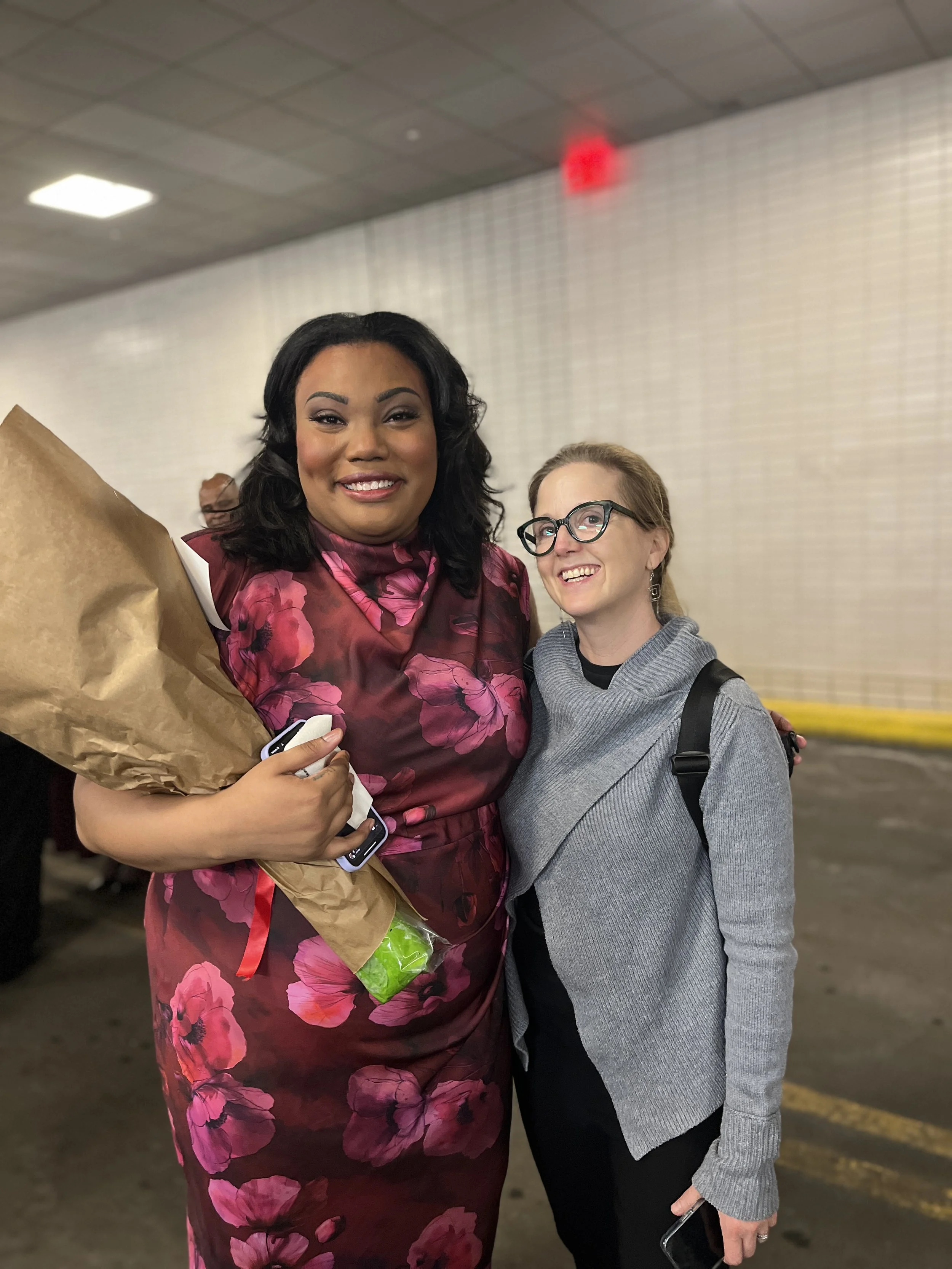 Two women standing together indoors, smiling at the camera. The woman on the left is holding a bouquet of flowers wrapped in brown paper and has a phone in her hand. The woman on the right is wearing glasses and a gray sweater and is carrying a backp