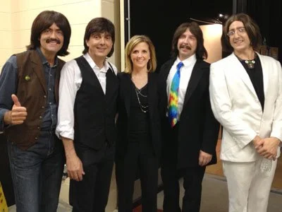 Five people standing together in a group, smiling and dressed in casual and formal attire, with a black backdrop and studio lighting.