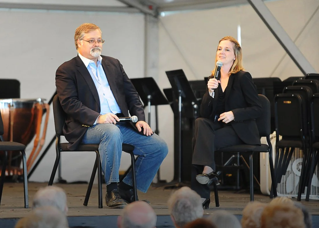 A man and a woman sitting on chairs on a stage, engaged in a discussion. The man, wearing glasses, a suit jacket, and jeans, is holding a microphone. The woman, dressed in a black suit, is speaking into a microphone. In the background, there are musi