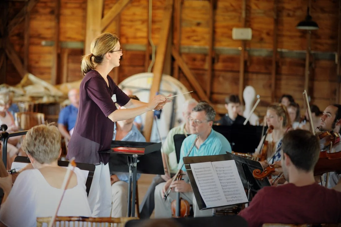 Erin Freeman conducting an orchestra in a rustic wooden venue, with musicians playing string instruments and reading sheet music.