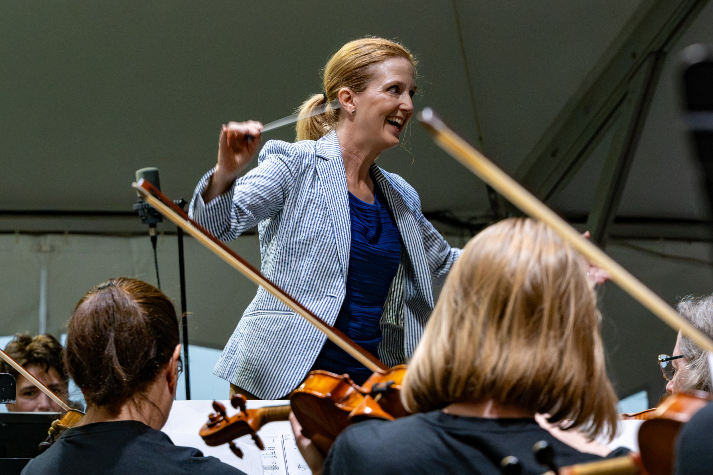 Erin Freeman conducting an orchestra with a smiling expression, holding a baton, while orchestra members play violins in an outdoor tent.