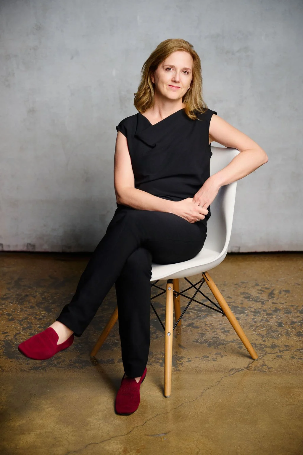 A woman with shoulder-length reddish-brown hair sitting on a white chair with wooden legs, wearing a black sleeveless top, black pants, and red shoes, against a gray textured wall.