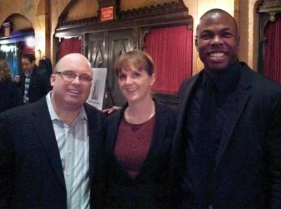 Three people smiling inside a restaurant or banquet hall with wood paneling and red curtains in the background.
