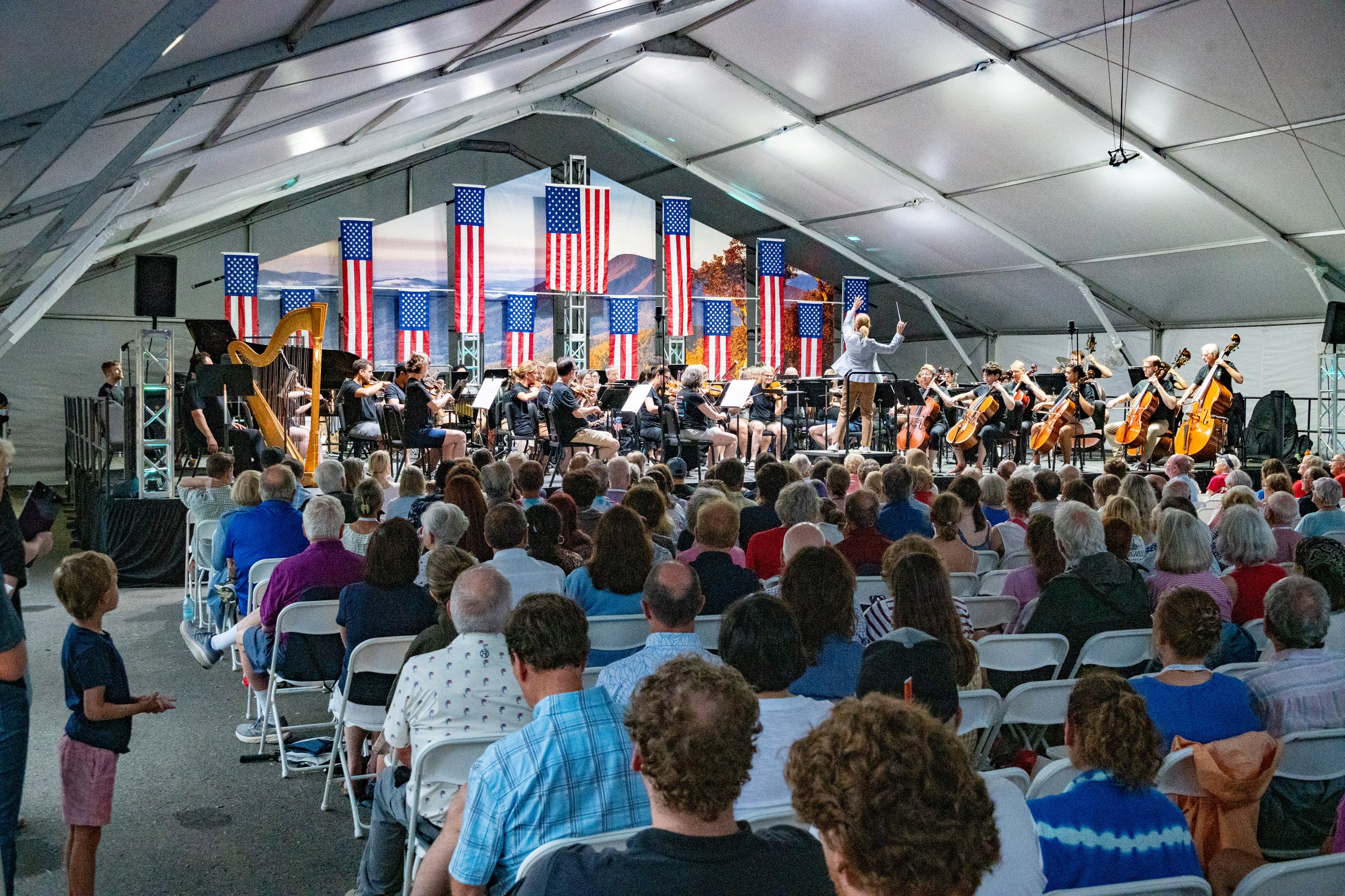 Erin Freeman conducting an orchestra performing on stage inside a large tent, decorated with American flags and a mountain landscape backdrop, with an audience seated in front.