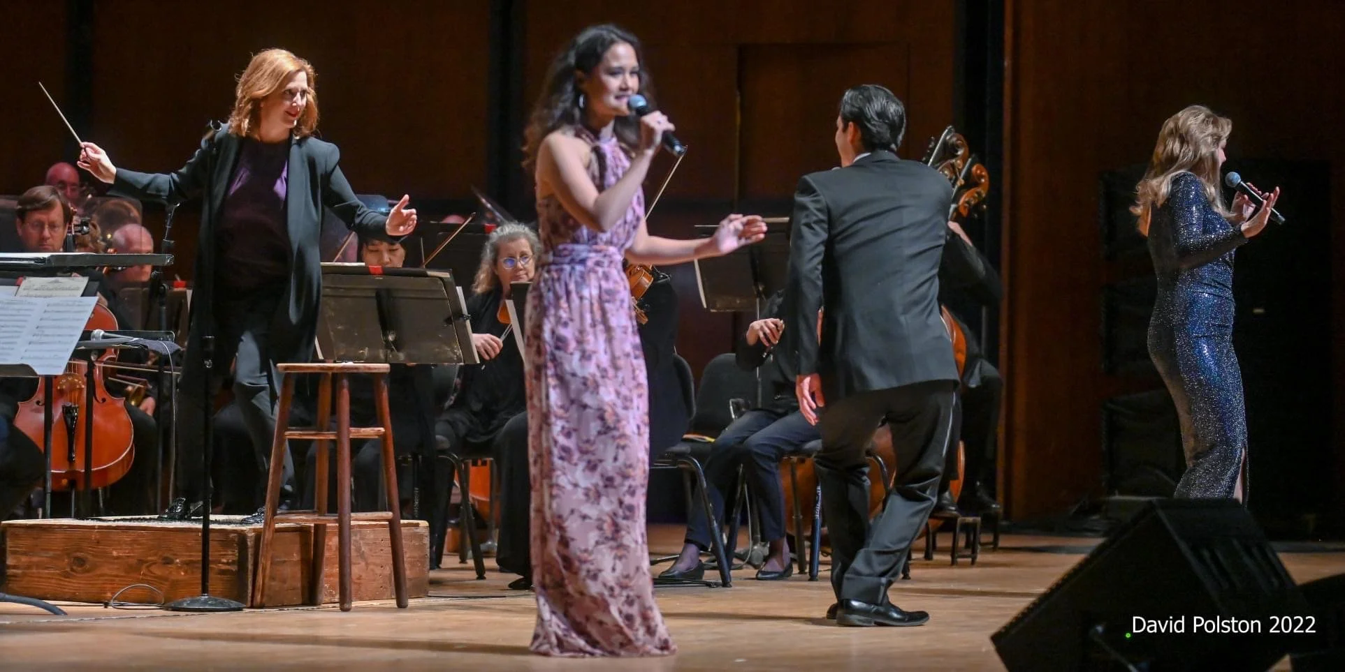 Photo of a live musical performance featuring three female singers on stage with musicians and orchestral instruments in the background.