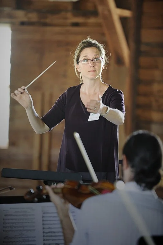 A woman conducting an orchestra with a music stand and a violinist in the foreground. She is holding a baton and appears to be giving directions to the musicians in a wooden-paneled room.