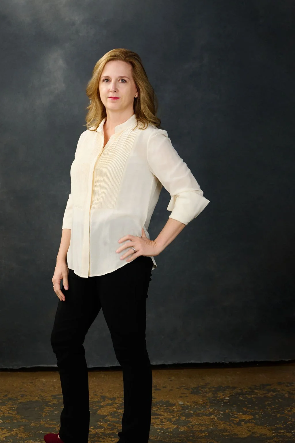 Erin Freeman standing against a dark gray background, wearing a cream-colored blouse and black pants, with her right hand on her hip.