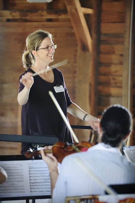 A woman conducting an orchestra with a violinist in the foreground, both in a rustic wooden venue.