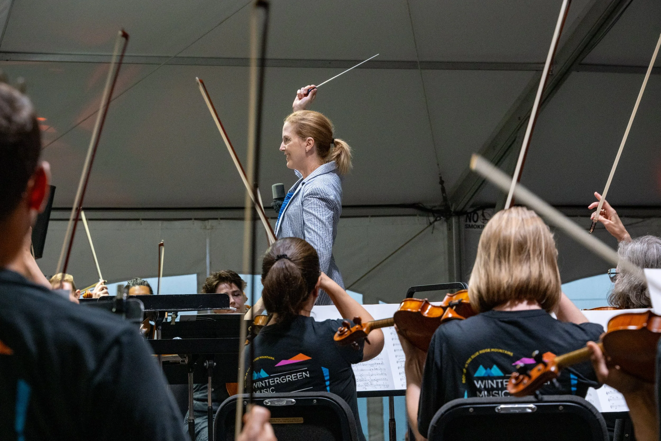 An orchestra with musicians playing violins and a conductor leading the group, inside a large tent with a gray ceiling.