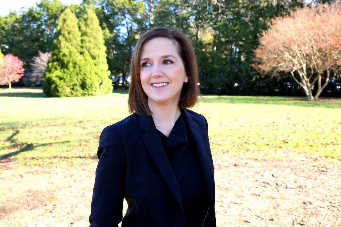 Erin Freeman wearing a black blazer, smiling while standing outdoors in a park with colorful trees and green grass during autumn.