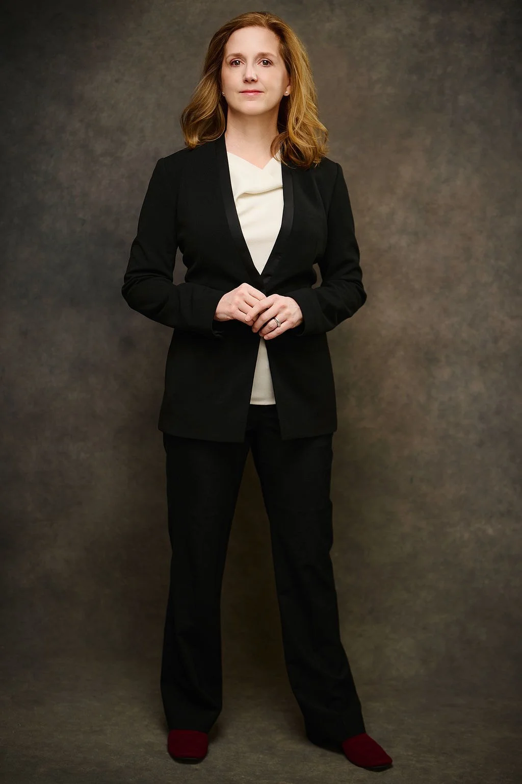 Erin Freeman standing against a neutral background, wearing a black blazer, cream-colored blouse, black pants, and red shoes.