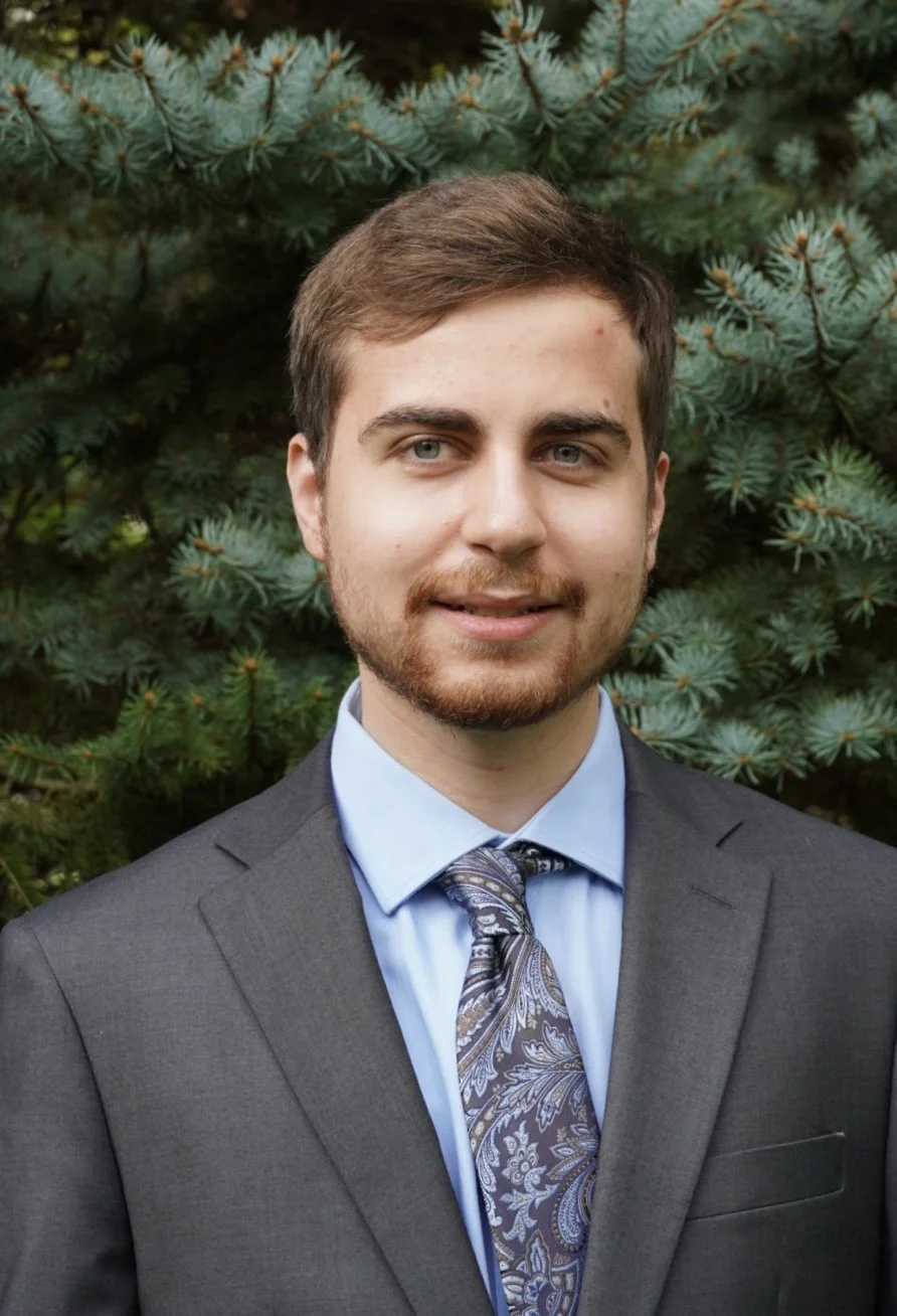A young man with light brown hair and a beard wearing a gray suit, light blue shirt, and patterned tie, standing outdoors in front of green pine trees.