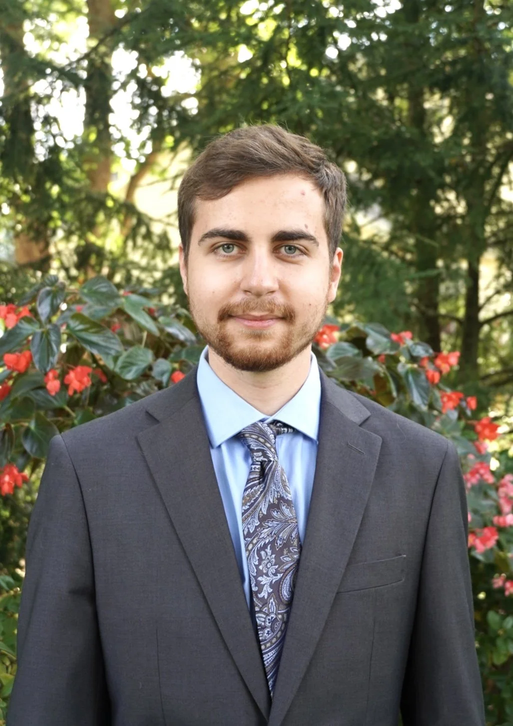 A young man with brown hair, blue eyes, and a beard dressed in a gray suit, light blue shirt, and a paisley tie, standing outdoors with greenery and red flowers in the background.