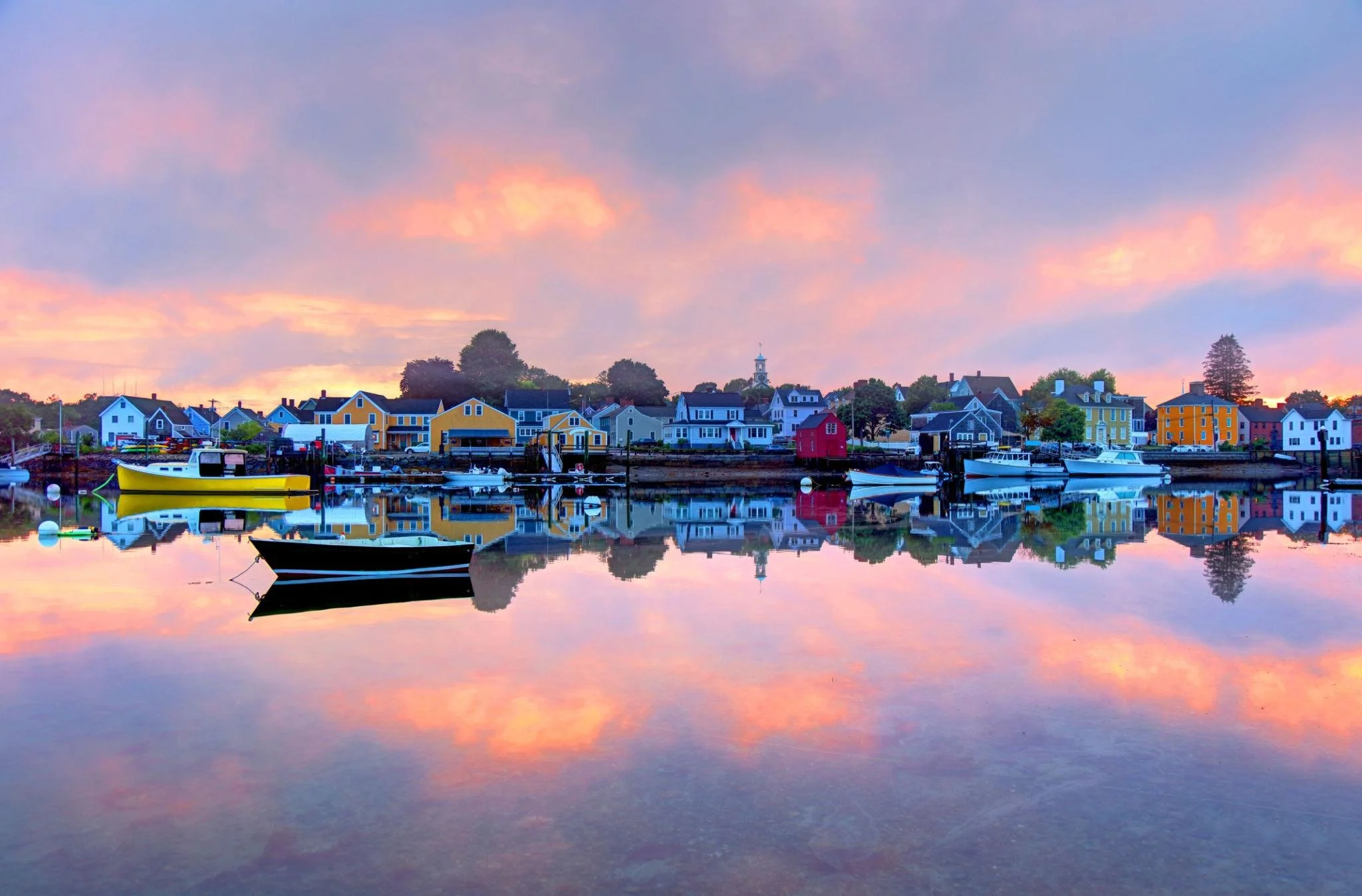 Colorful lakeside village at sunset with colorful houses, boats docked in calm water, and a pink, purple, and blue sky reflected on the water.