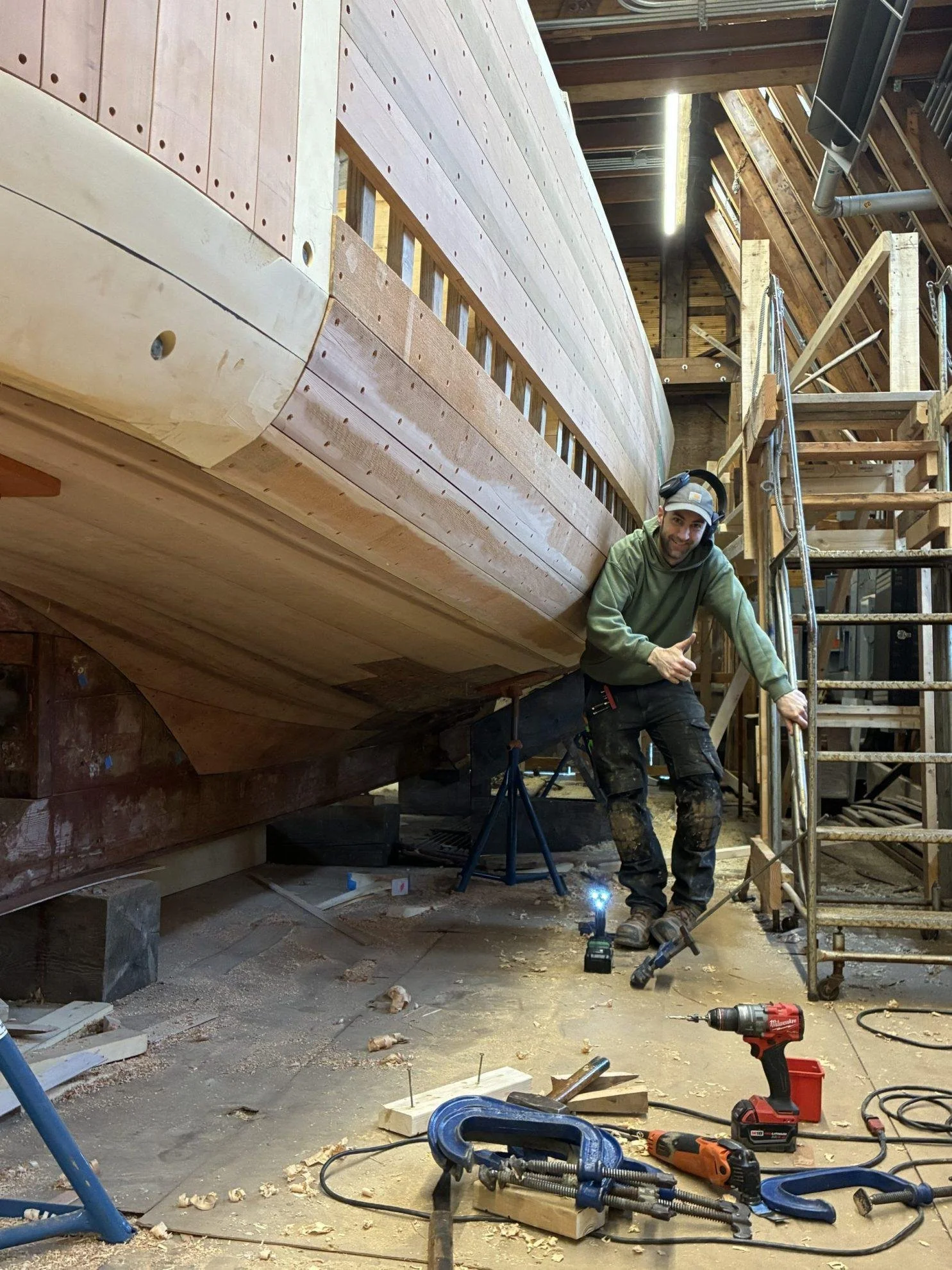 A man working on the construction of a wooden boat in a workshop, surrounded by tools and scaffolding.
