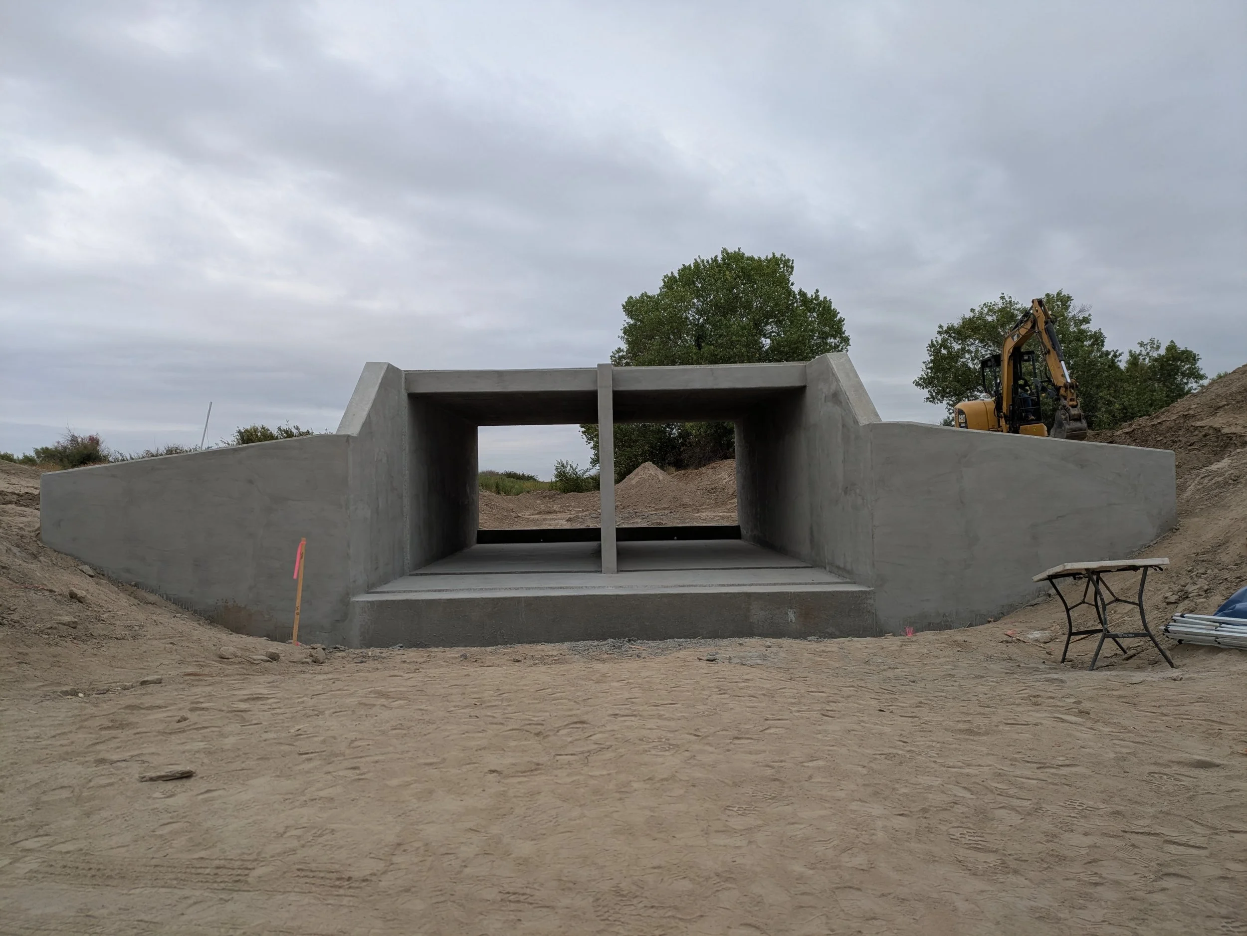 Construction site with a concrete structure, a yellow excavator, and trees under a cloudy sky.