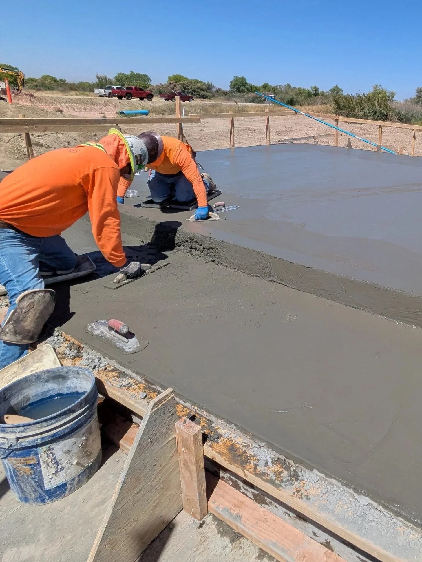 Two construction workers are kneeling on a freshly poured concrete slab, leveling the surface with trowels at a construction site. They are wearing orange shirts, gloves, and helmets. Construction materials and a bucket filled with water or concrete are seen nearby, with a backdrop of parked trucks and a clear blue sky.