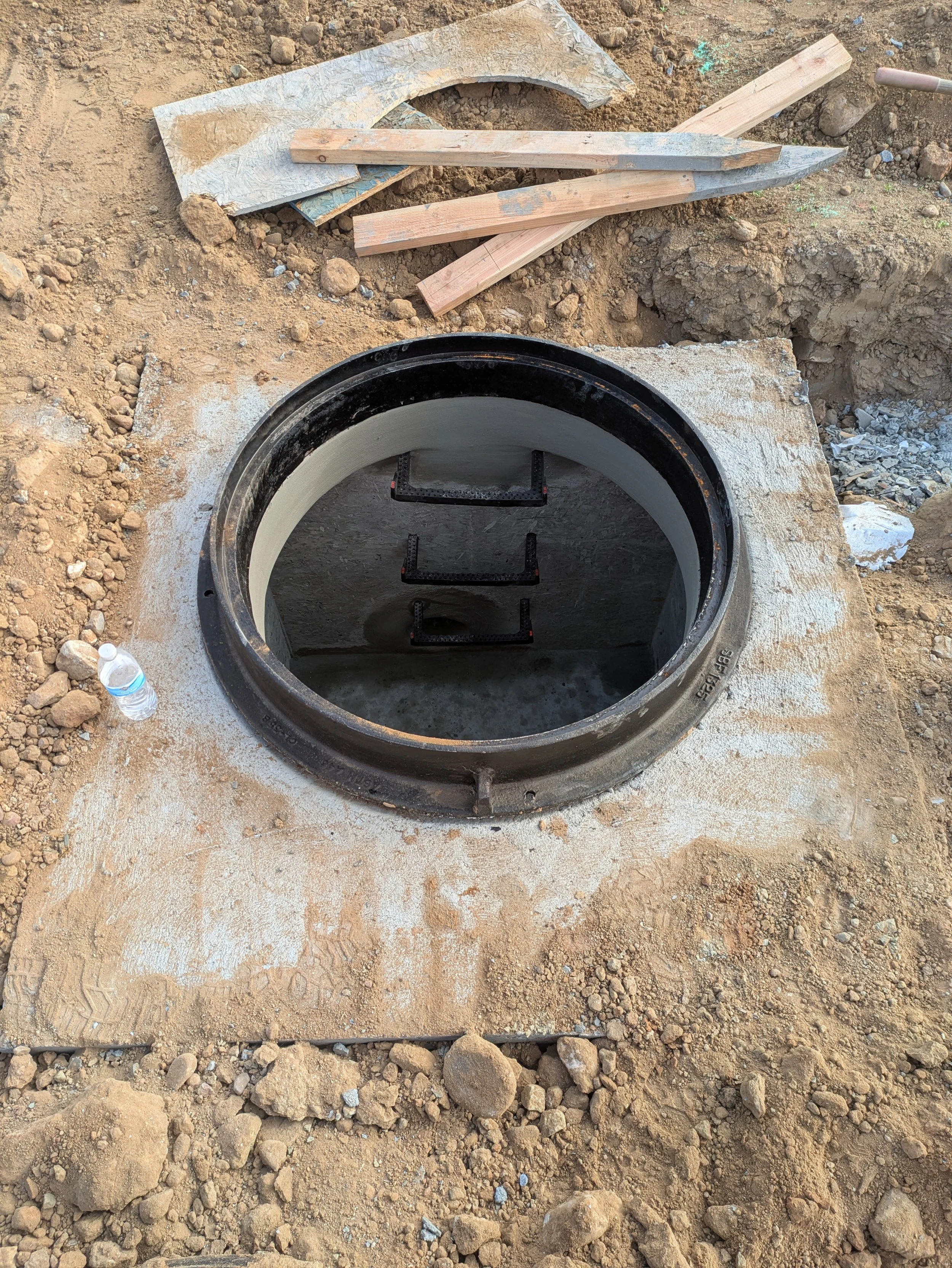 Construction site with a partially dug hole, concrete, and wooden planks around it. A black pipe is embedded into the ground, and a plastic water bottle is on the dirt surface.