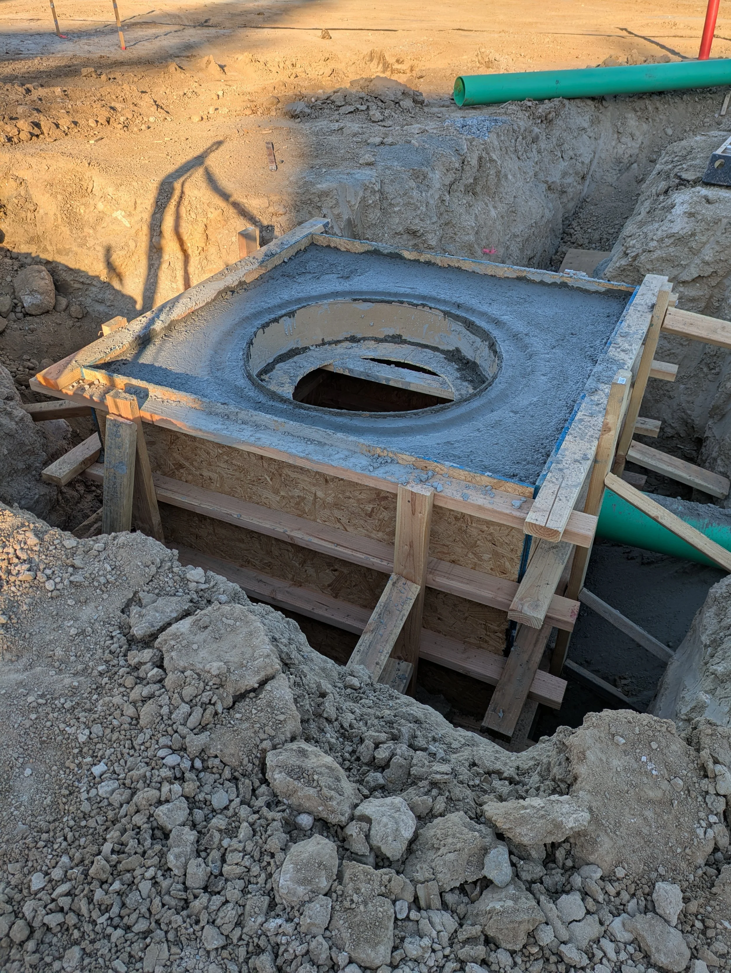 Construction site with excavation trench, wooden formwork, concrete, and a green pipe, surrounded by dirt and rocks.