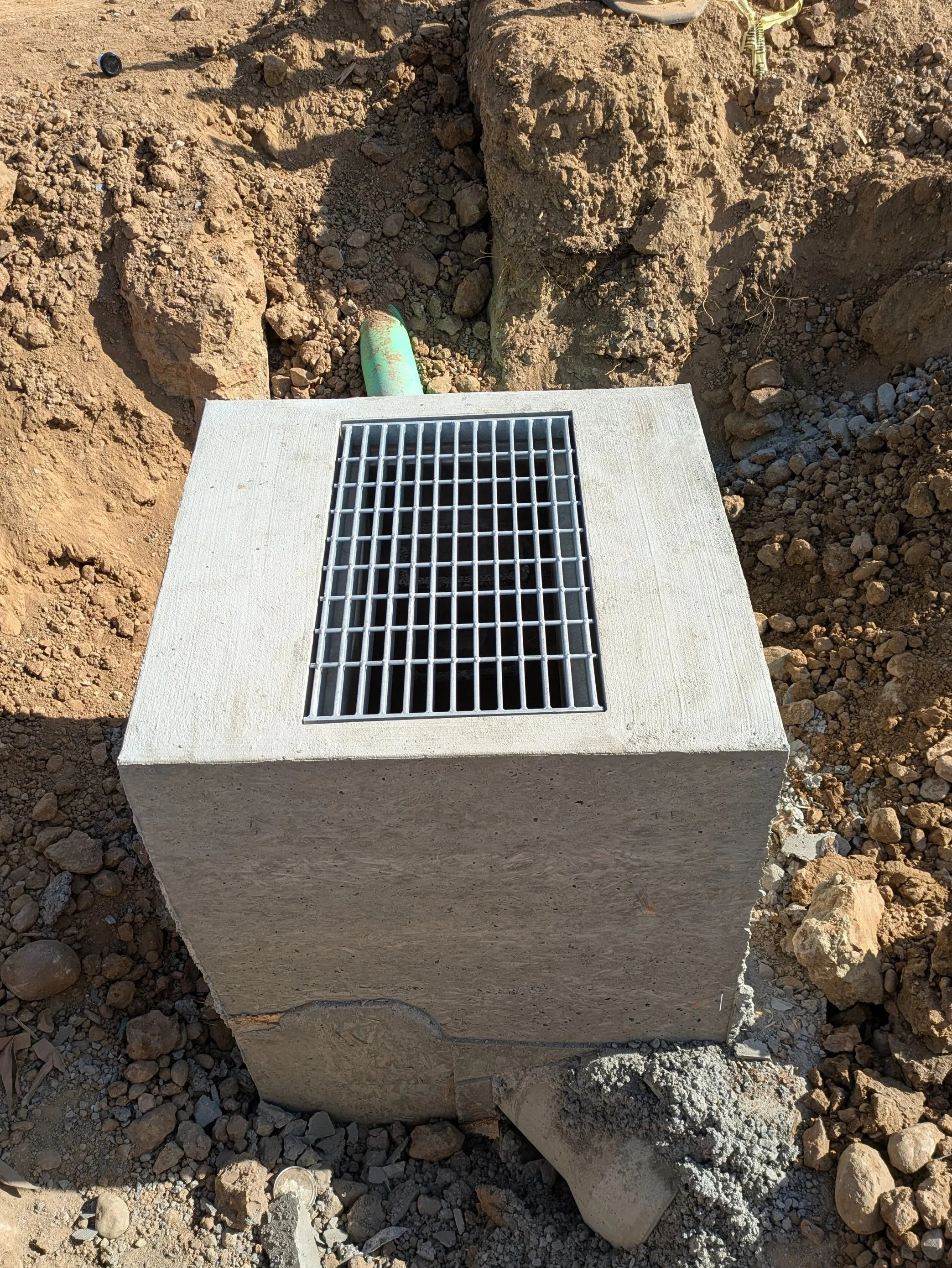 A concrete structure with a metal grate on top, surrounded by dirt and rocks, with a green pipe visible underneath.