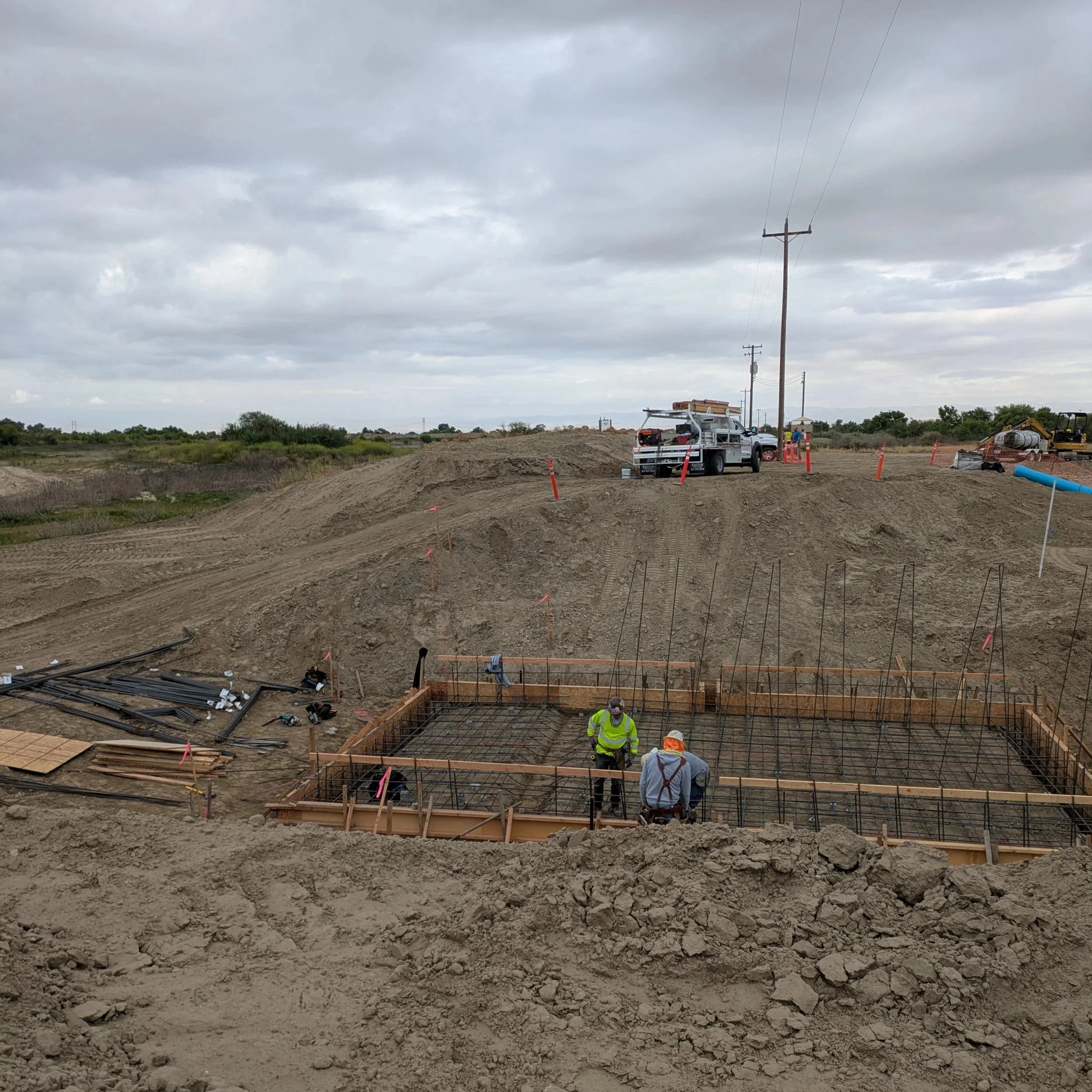 Construction workers at a building site with a foundation frame and rebar, surrounded by equipment and vehicles, under cloudy skies.