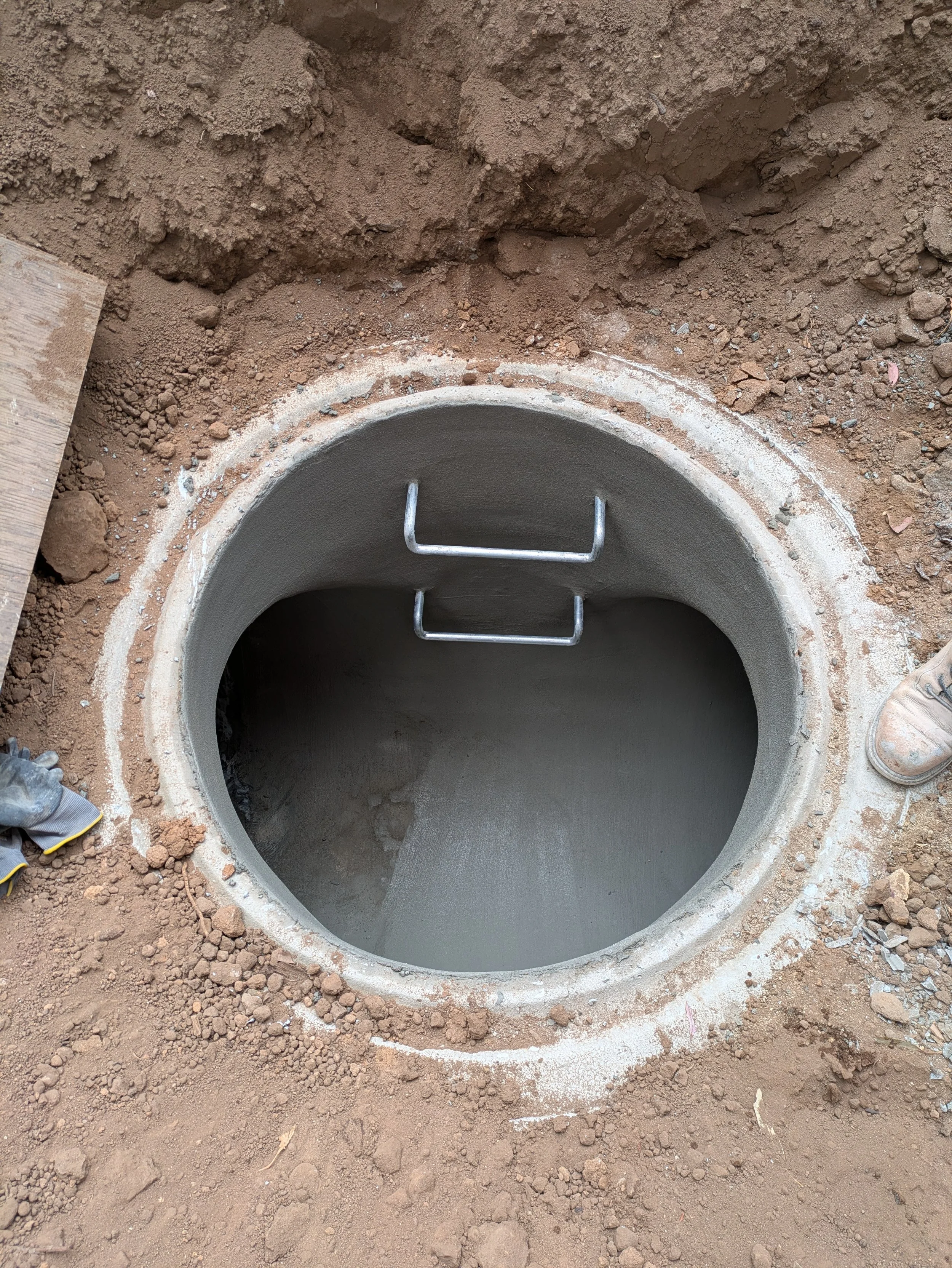 Concrete manhole with a metallic ladder inside, surrounded by dirt and gravel.