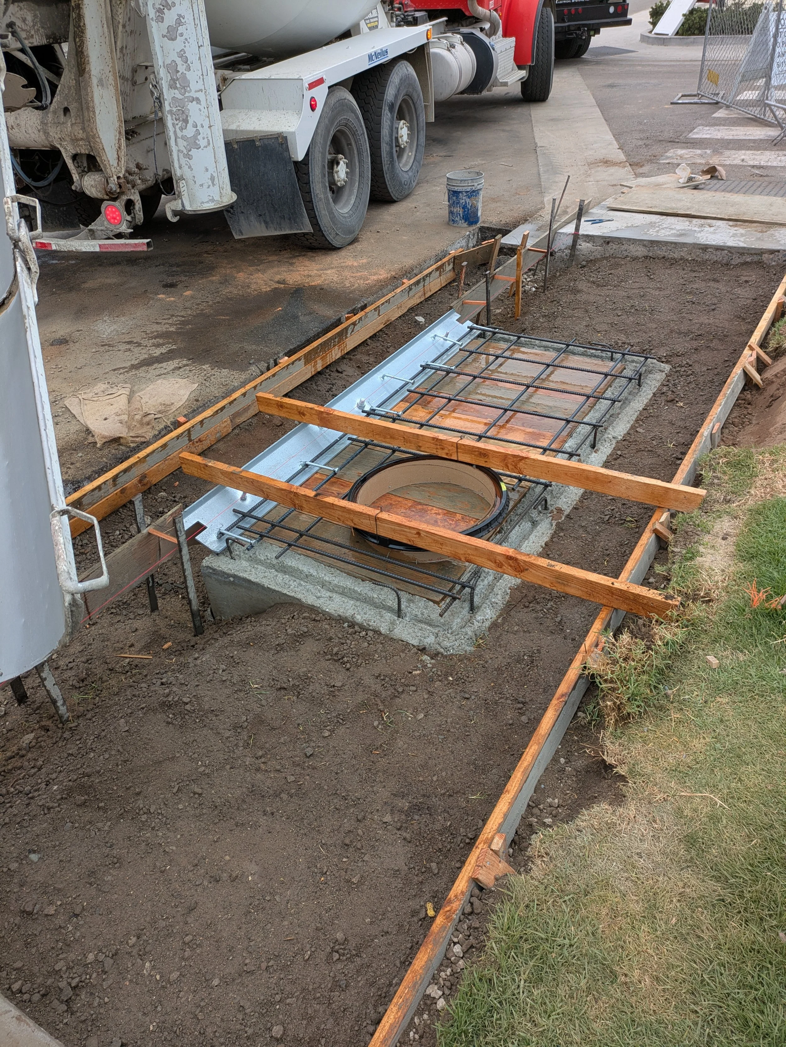 Construction site with a concrete foundation in progress, surrounded by wooden and metal framing, a construction wheelbarrow, and a large truck in the background.