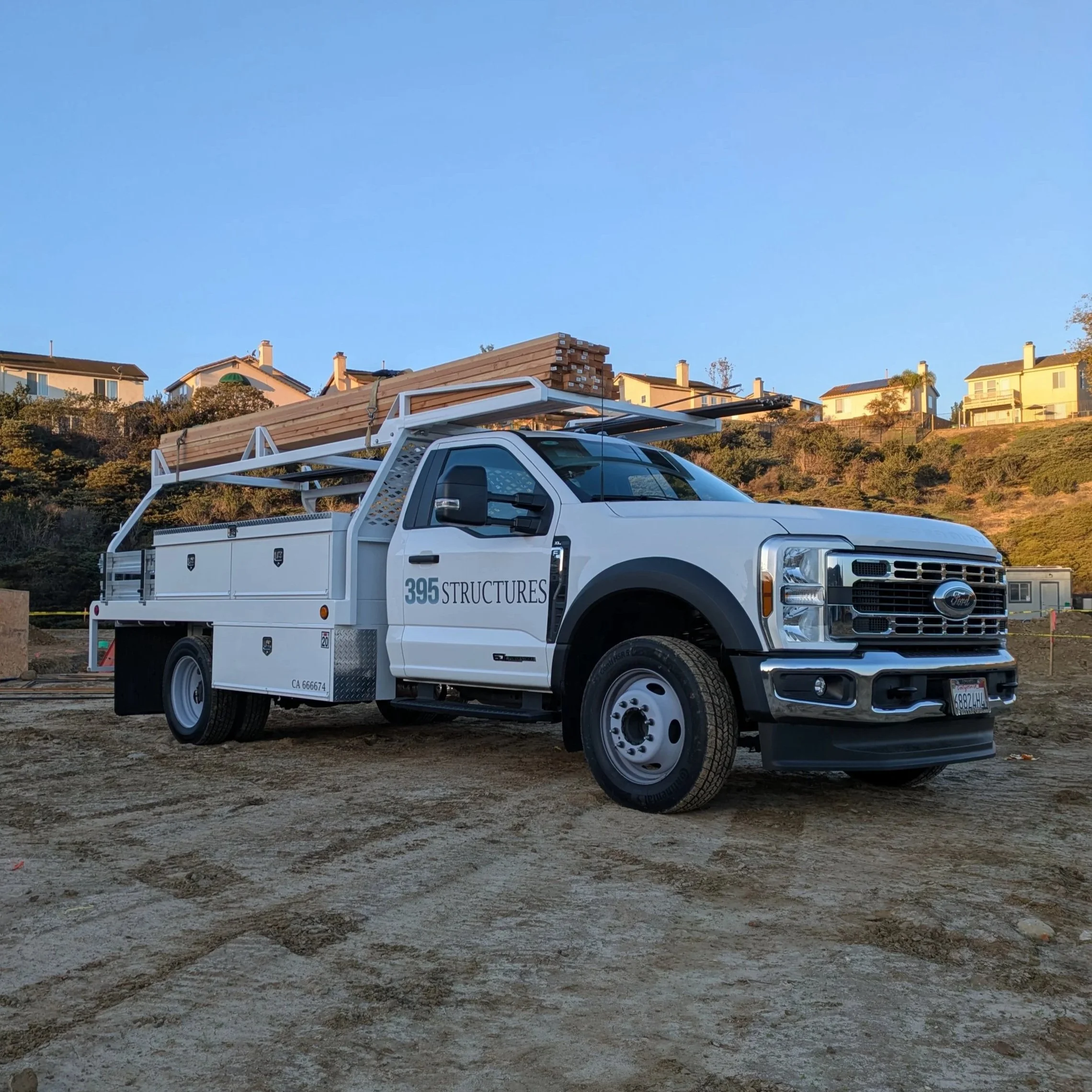 A white pickup truck with a ladder rack carrying long wooden planks parked on a dirt or gravel lot. In the background, there are residential houses on a hillside with trees and bushes.