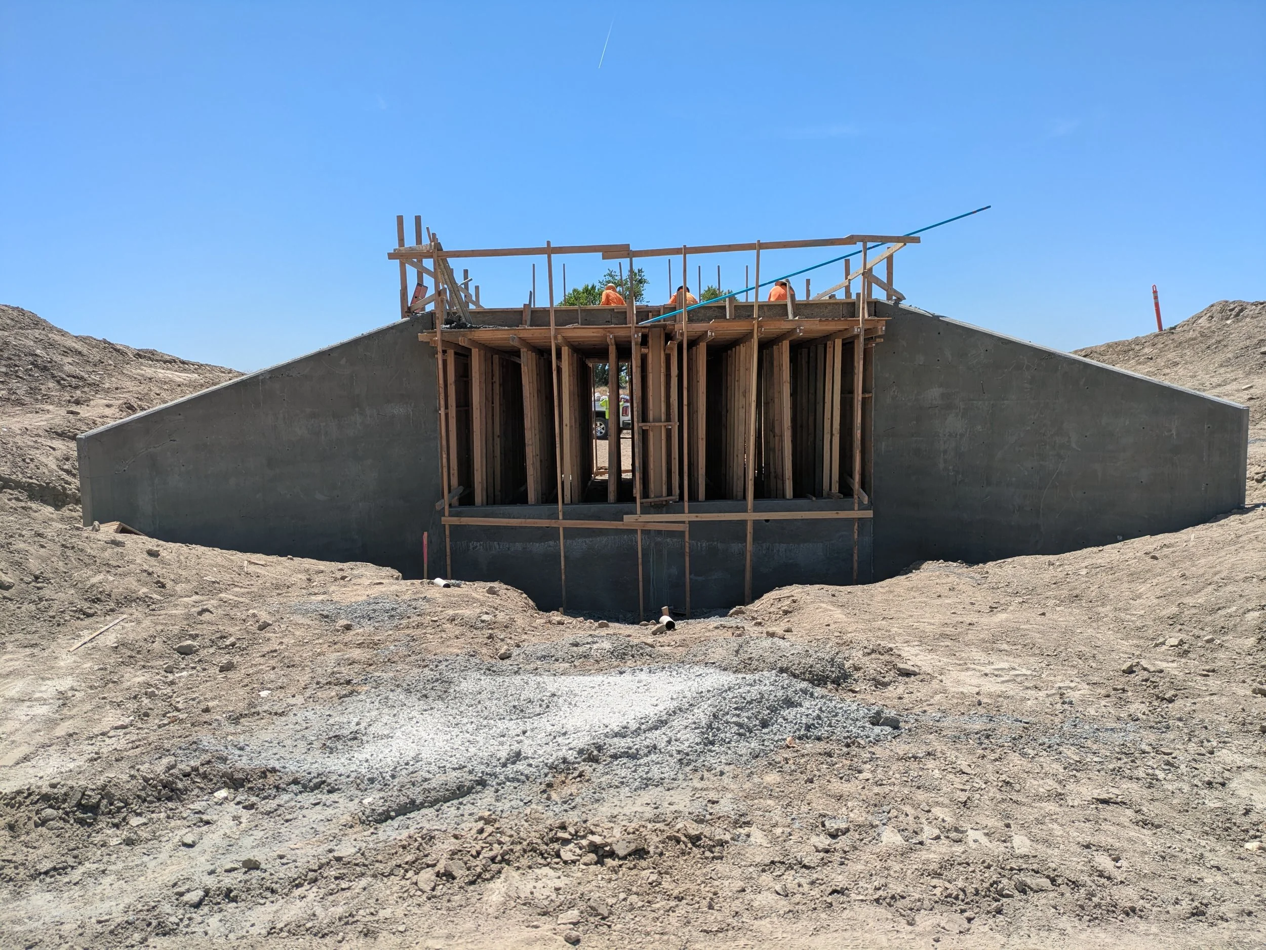 Construction site with a bridge or culvert under construction, workers in orange safety vests working on the structure, and a blue sky overhead.